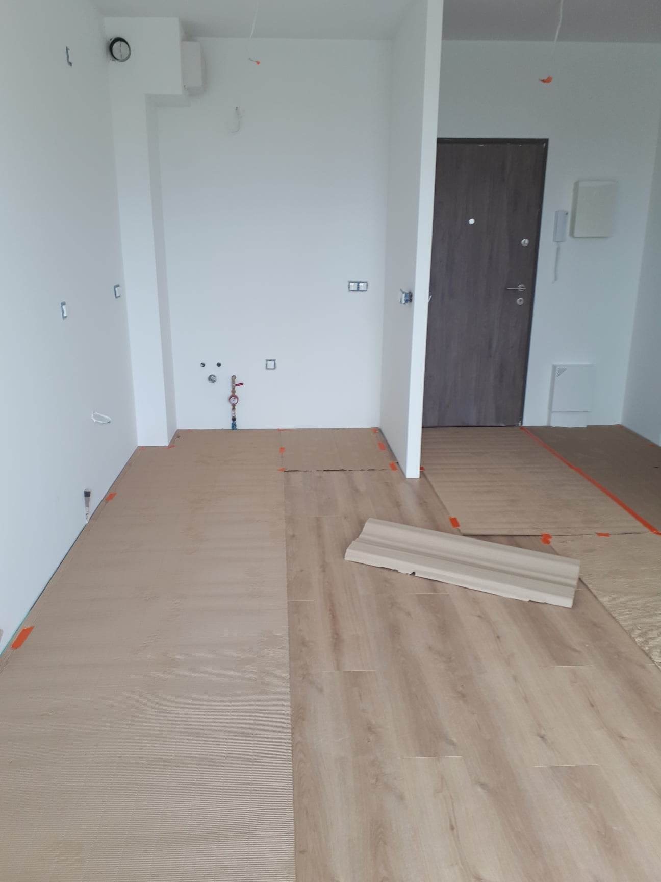 Układanie paneli podłogowych w jasnym wnętrzu, widoczne przygotowane podłoże, fragment listwy przypodłogowej i instalacja wodna na ścianie.