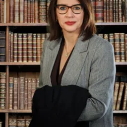 Kobieta w okularach i szarej marynarce, trzymająca czarną marynarkę na tle regału z książkami.