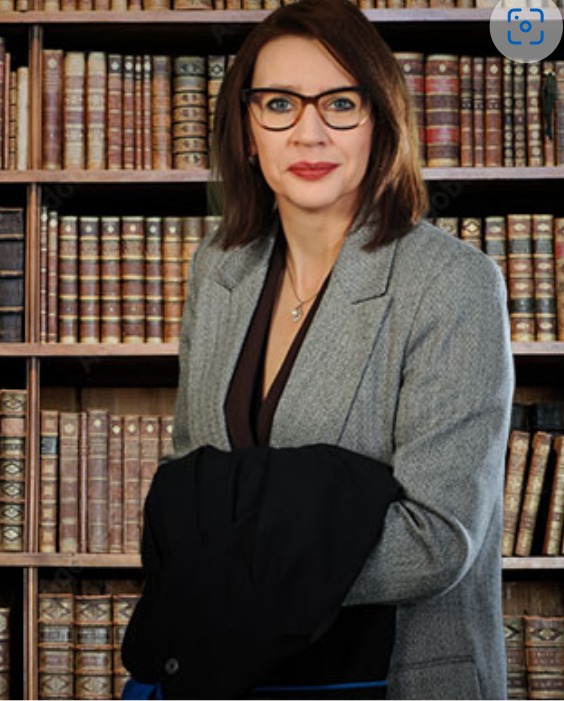 Kobieta w okularach i szarej marynarce, trzymająca czarną marynarkę na tle regału z książkami.