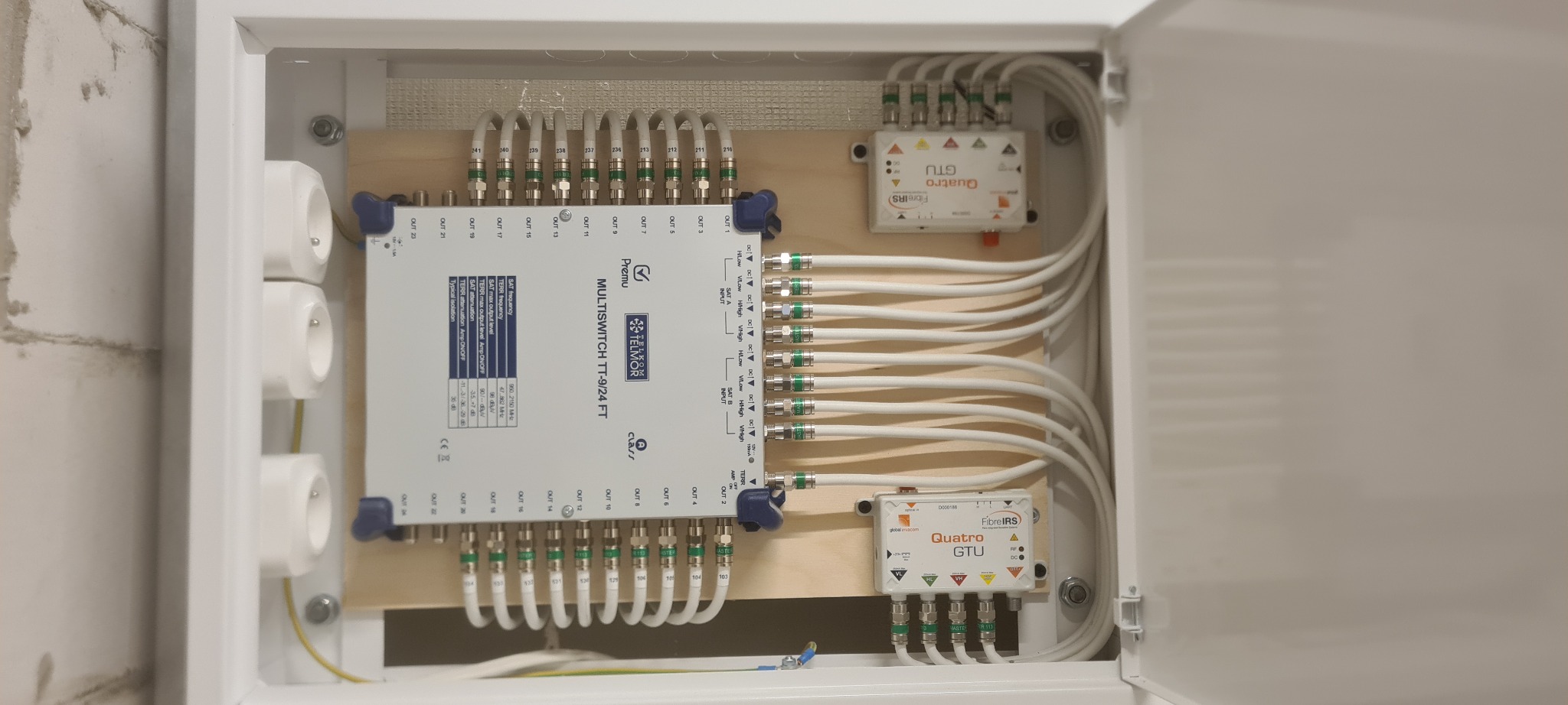 Rozdzielnica telekomunikacyjna w białej obudowie, z multiswitchem Premu Telmor TT-9/24 FT, modułami Quatro GTU FibreIRS i licznymi kablami koncentrycznymi, zamontowana na drewnianej płycie...