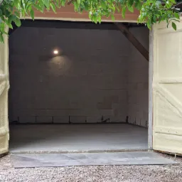 Otwarty garaż z betonową podłogą i ścianami z pustak&oacute;w, ocieplony pianką PUR na drzwiach i framudze, widoczne gałęzie drzewa w g&oacute;rnej części kadru.