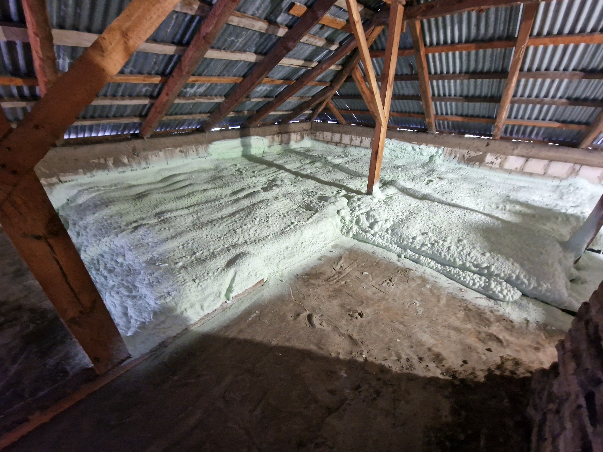 Poddasze z widoczną warstwą jasnej pianki izolacyjnej, pokrywającej ściany i podłogę, drewniana konstrukcja dachu z blachą falistą w tle.