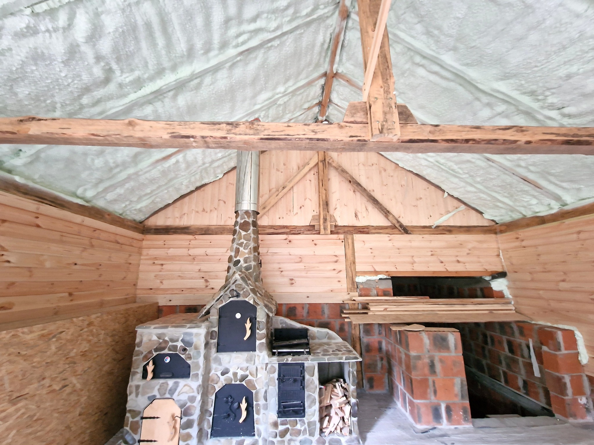 Poddasze z drewnianą konstrukcją i kamiennym piecem, ocieplone pianką PUR, widoczne ślady pianki na dachu i elementach konstrukcyjnych.