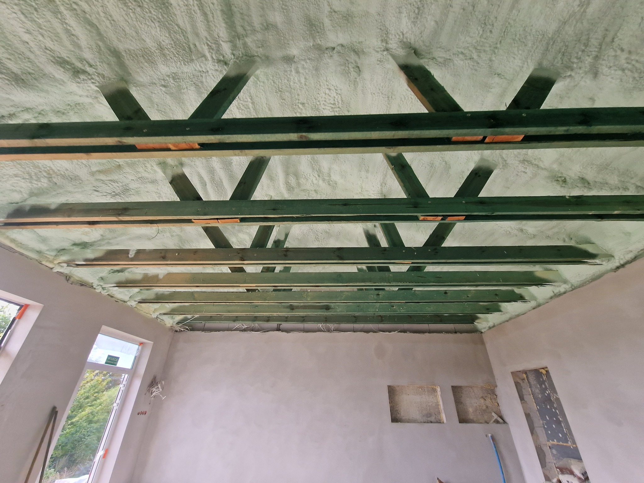 Poddasze z drewnianą konstrukcją pokryte białą pianką izolacyjną, widoczne okna i surowe ściany w trakcie wykańczania.