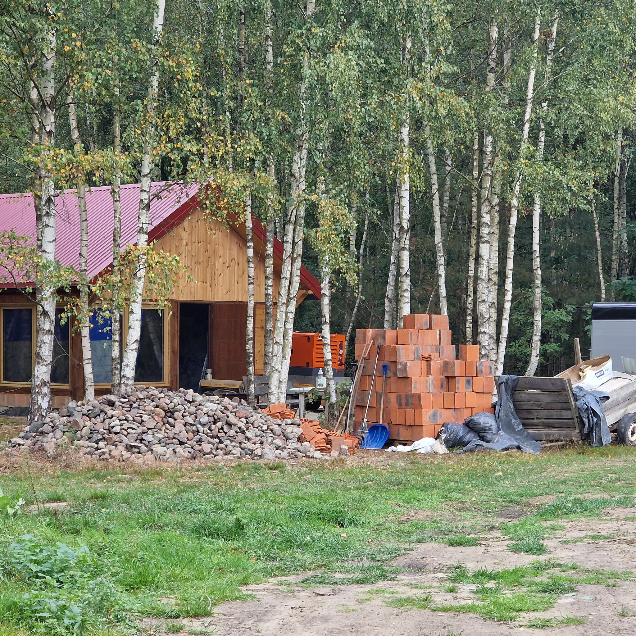Budowa domu z drewnianą elewacją i czerwonym dachem, widoczny stos kamieni, paleta z cegłami, narzędzia budowlane i przyczepka, otoczenie stanowią brzozy i trawa.