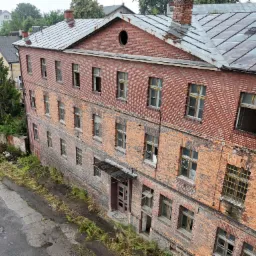 Trzykondygnacyjny, ceglany budynek z metalowym dachem widziany z góry. Widoczne uszkodzenia elewacji, powybijane okna i zarośnięte otoczenie. Na dachu komin i okrągłe okno.
