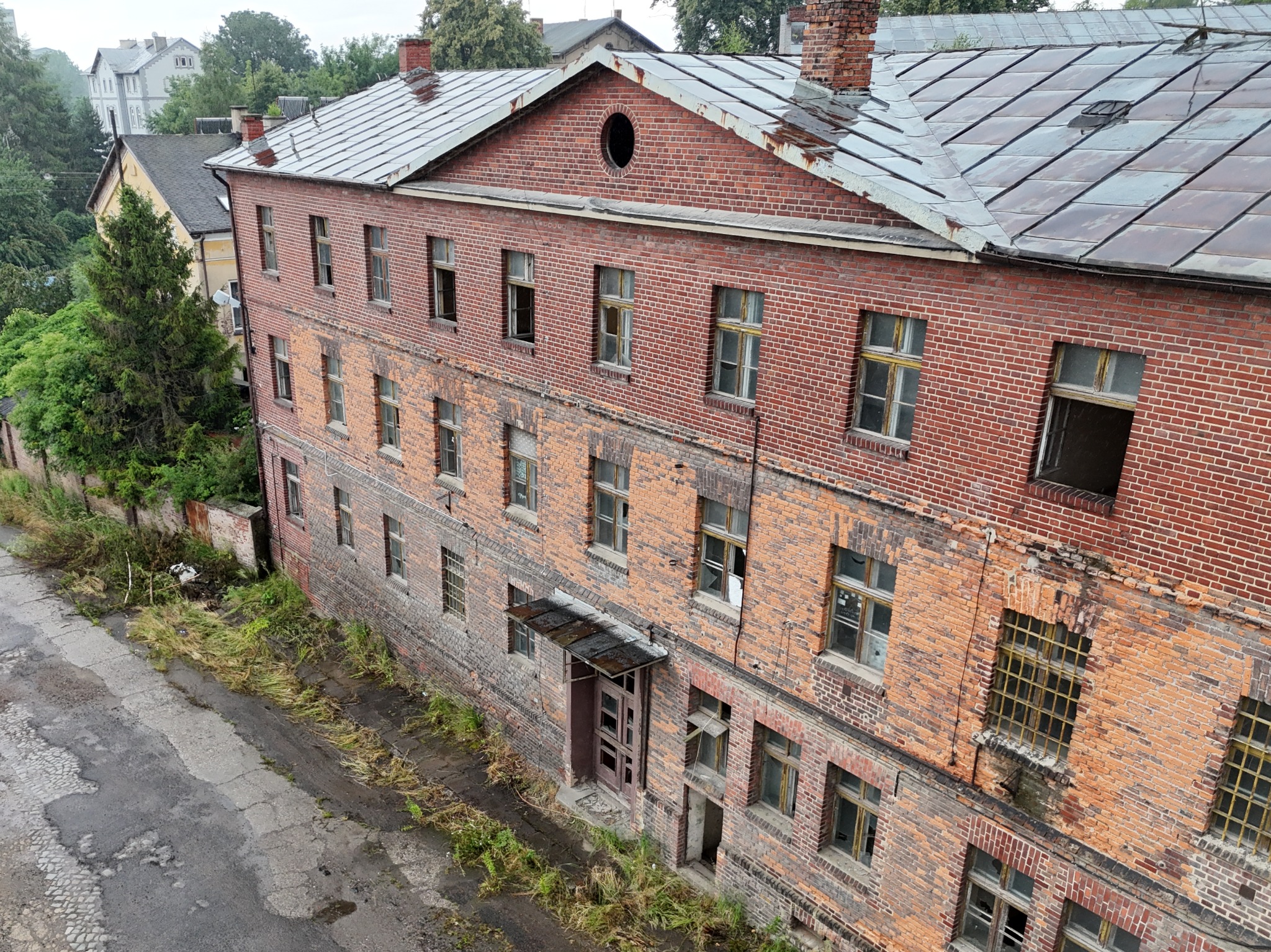 Trzykondygnacyjny, ceglany budynek z metalowym dachem widziany z góry. Widoczne uszkodzenia elewacji, powybijane okna i zarośnięte otoczenie. Na dachu komin i okrągłe okno.