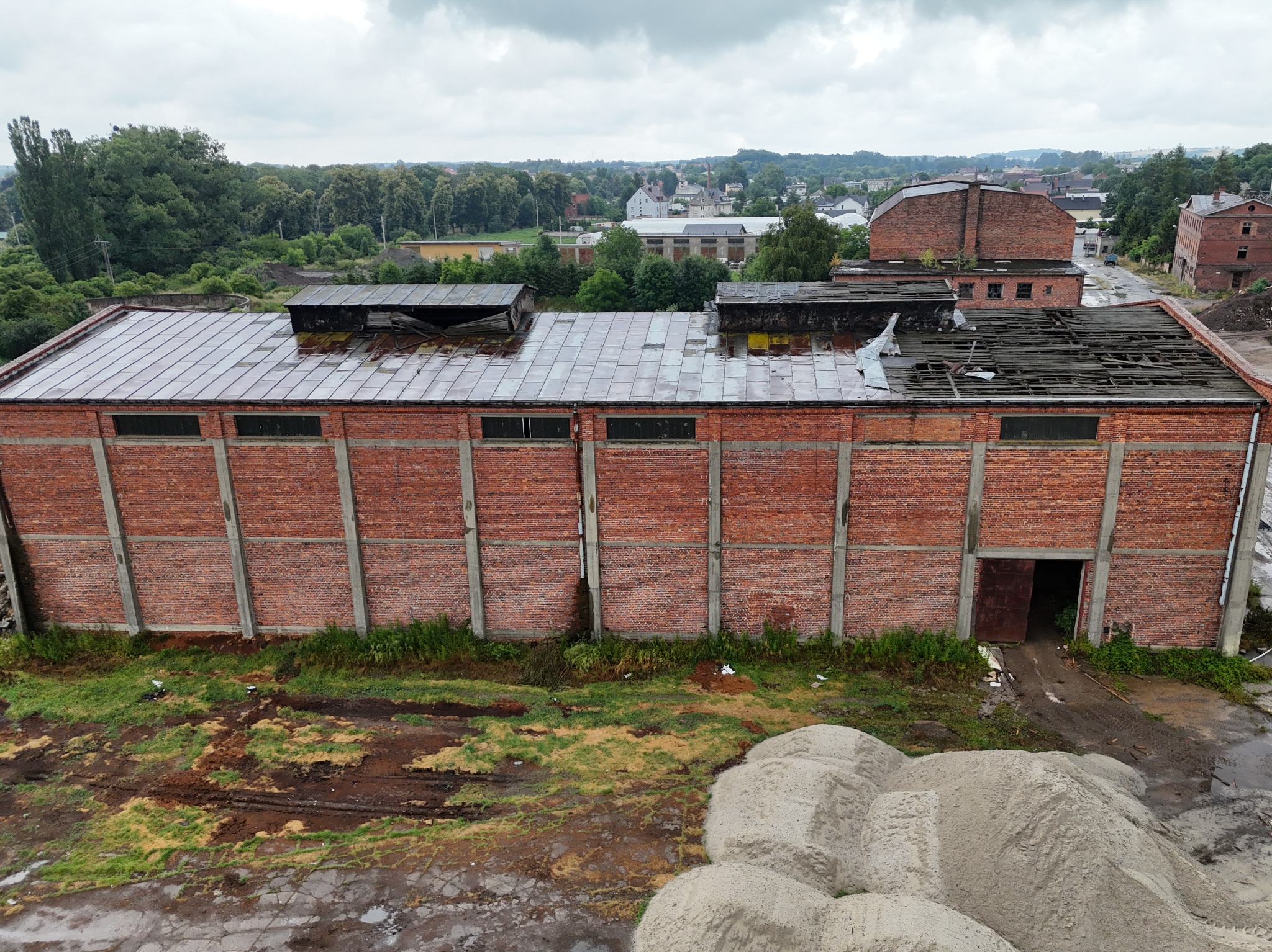 Zniszczony ceglany budynek magazynowy z dziurawym dachem i otwartą bramą, widok z góry na hałdy piasku i zarośnięte tereny.
