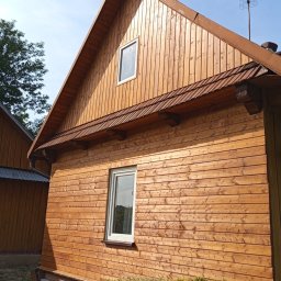 Altanki tarasy i meble ogrodowe - Drewniany dom z jasną elewacją, widoczny z boku, z małym oknem na poddaszu i anteną na dachu, otoczony zielenią. Świeżo pomalowana elewacja z widoczną strukturą drewna.