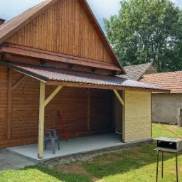 Drewniana konstrukcja z zadaszeniem przy budynku, częściowo obita deskami, z widocznym betonowym podłożem i narzędziami. W tle zieleń i budynek z czerwoną dachówką.