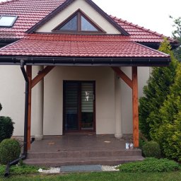 Altanki tarasy i meble ogrodowe - Zadaszony taras z drewnianą konstrukcją i ceramiczną dachówką, przylegający do domu z czerwonym dachem. Widoczne tuje i trawnik. Detal: ozdobne zakończenia słupów.