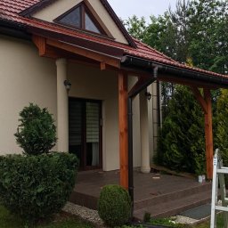 Altanki tarasy i meble ogrodowe - Zadaszenie tarasu z drewnianą konstrukcją i kolumnami, widok z ogrodu. Na trawniku leżą fragmenty płytek. Częściowo widoczna drabina.