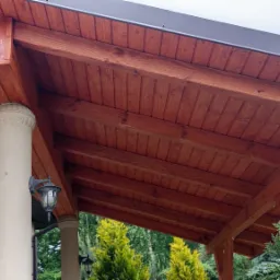 Zadaszenie tarasu z drewnianą konstrukcją i kolumną z oświetleniem, widok od dołu. Czerwono-brązowe drewno kontrastuje z zielenią drzew w tle.