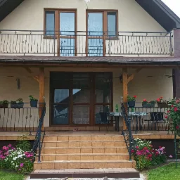 Dom z zadaszonym tarasem, balkonem z ozdobną balustradą, schodami wejściowymi i zielonym ogrodem z kwitnącymi różami.