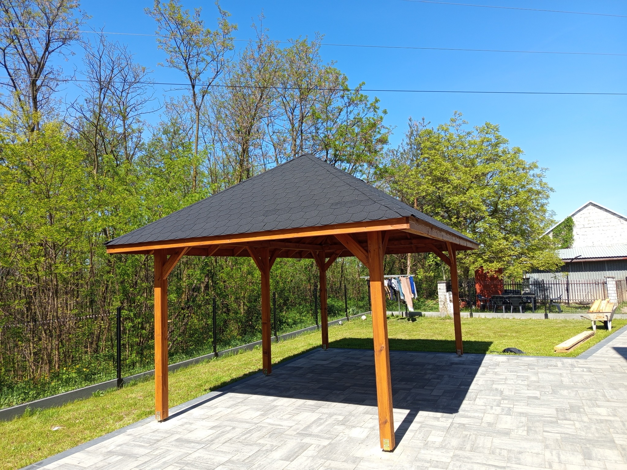 Drewniana altana z czarnym dachem na kostce brukowej, otoczona zielenią. W tle widoczny dom i suszarka na pranie. Słoneczny dzień, błękitne niebo.