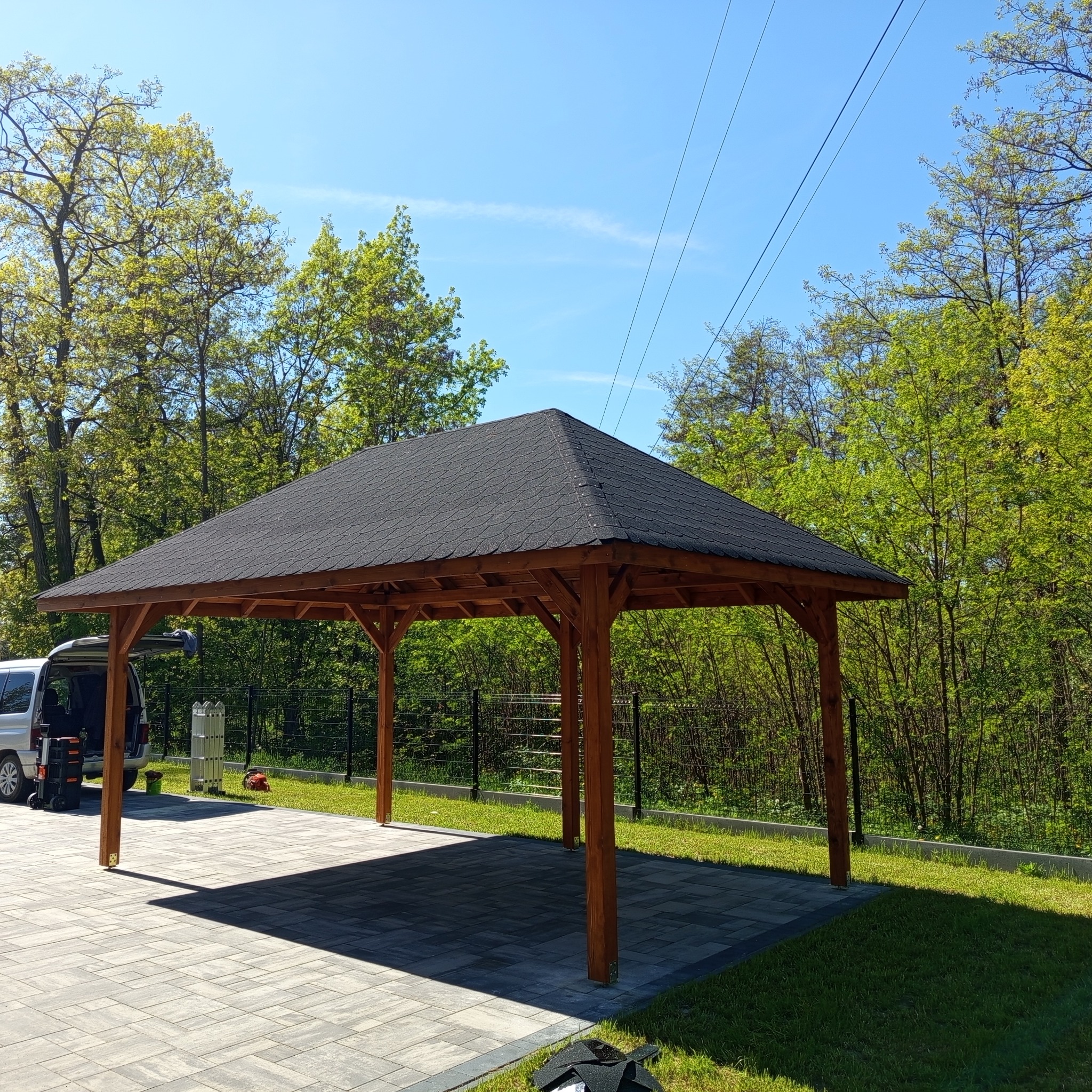 Zadaszona, drewniana konstrukcja z bitumicznym dachem na posesji. W tle zieleń drzew i błękitne niebo. Częściowo widoczny samochód dostawczy i narzędzia.