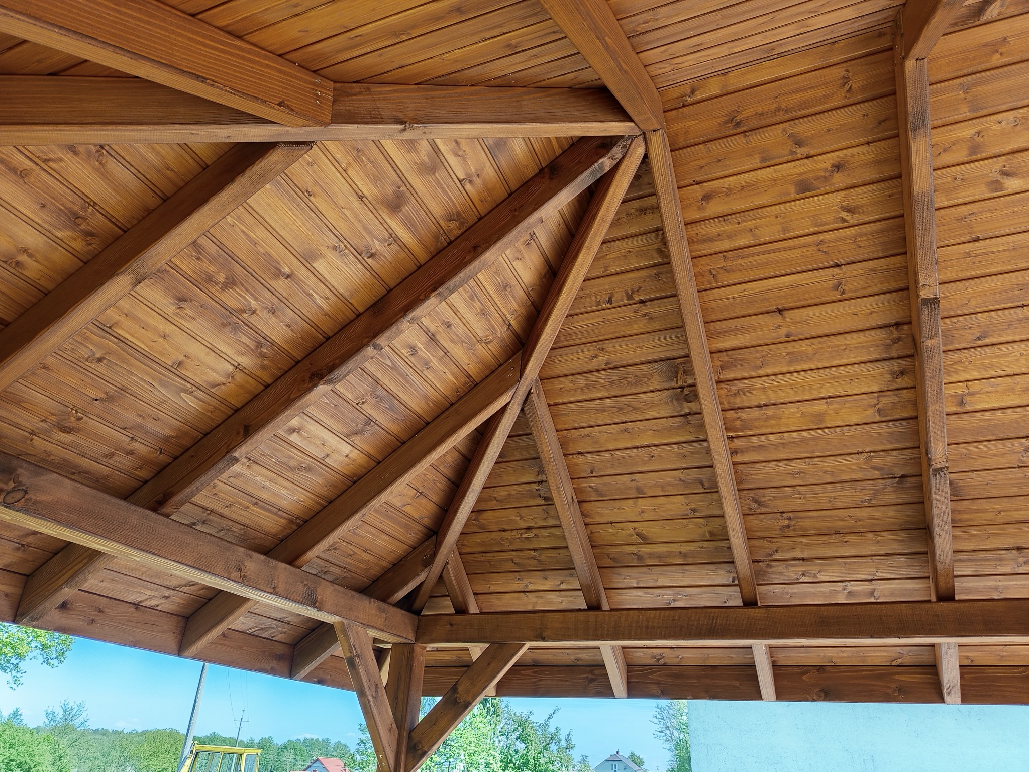 Drewniana konstrukcja dachu altany z widocznymi belkami i deskami w ciepłym odcieniu brązu, na tle błękitnego nieba i zieleni drzew. Widok z dołu.