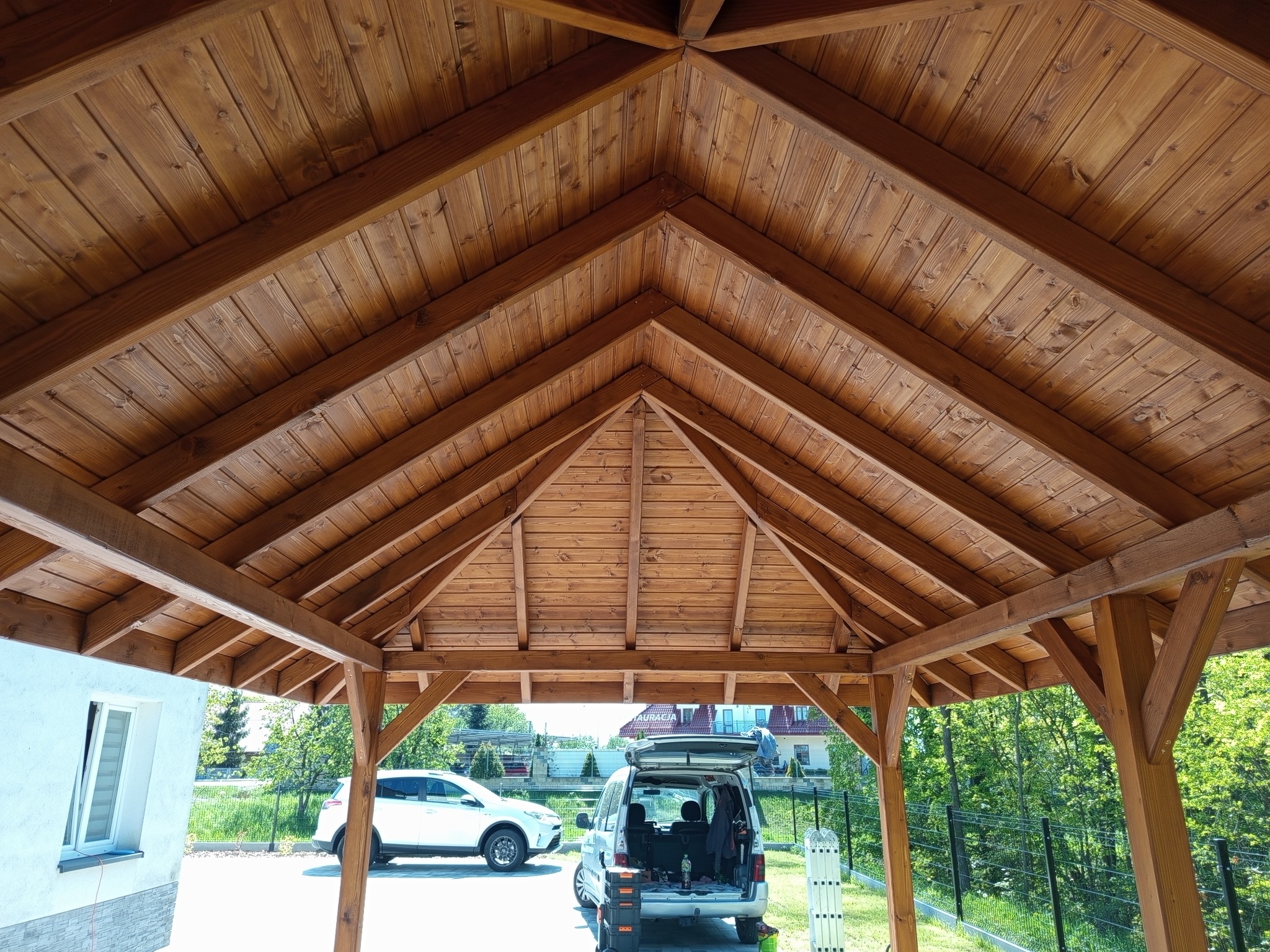 Drewniana altana z widoczną konstrukcją dachu z desek, w tle auta i zieleń. Widok od wewnątrz, z narzędziami w bagażniku auta. Ciepłe oświetlenie podkreśla strukturę drewna.