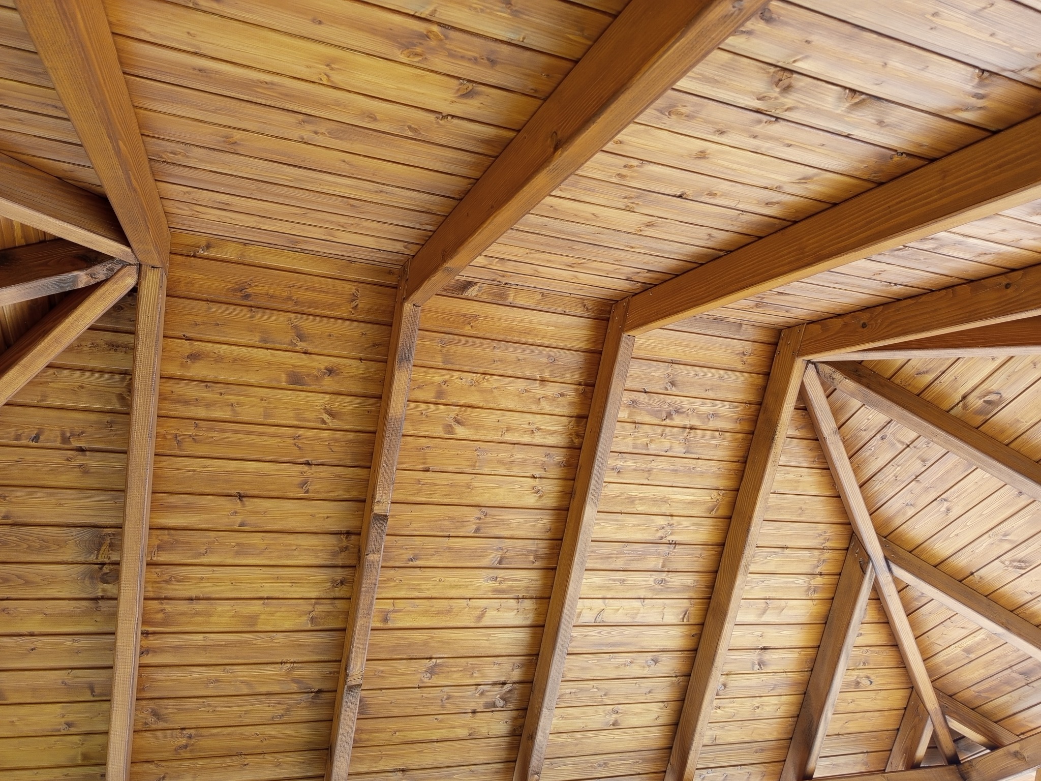 Drewniany sufit altany z widoczną konstrukcją więźby dachowej i deskowaniem. Naturalne odcienie drewna, ciepłe oświetlenie. Perspektywa z dołu.