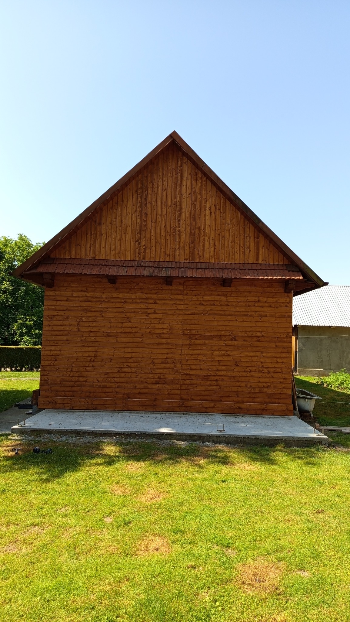 Drewniana konstrukcja z dachem dwuspadowym, wykończona poziomymi deskami, na betonowej podstawie. Widoczne elementy wykończeniowe dachu. Tło: trawnik i fragment budynku.