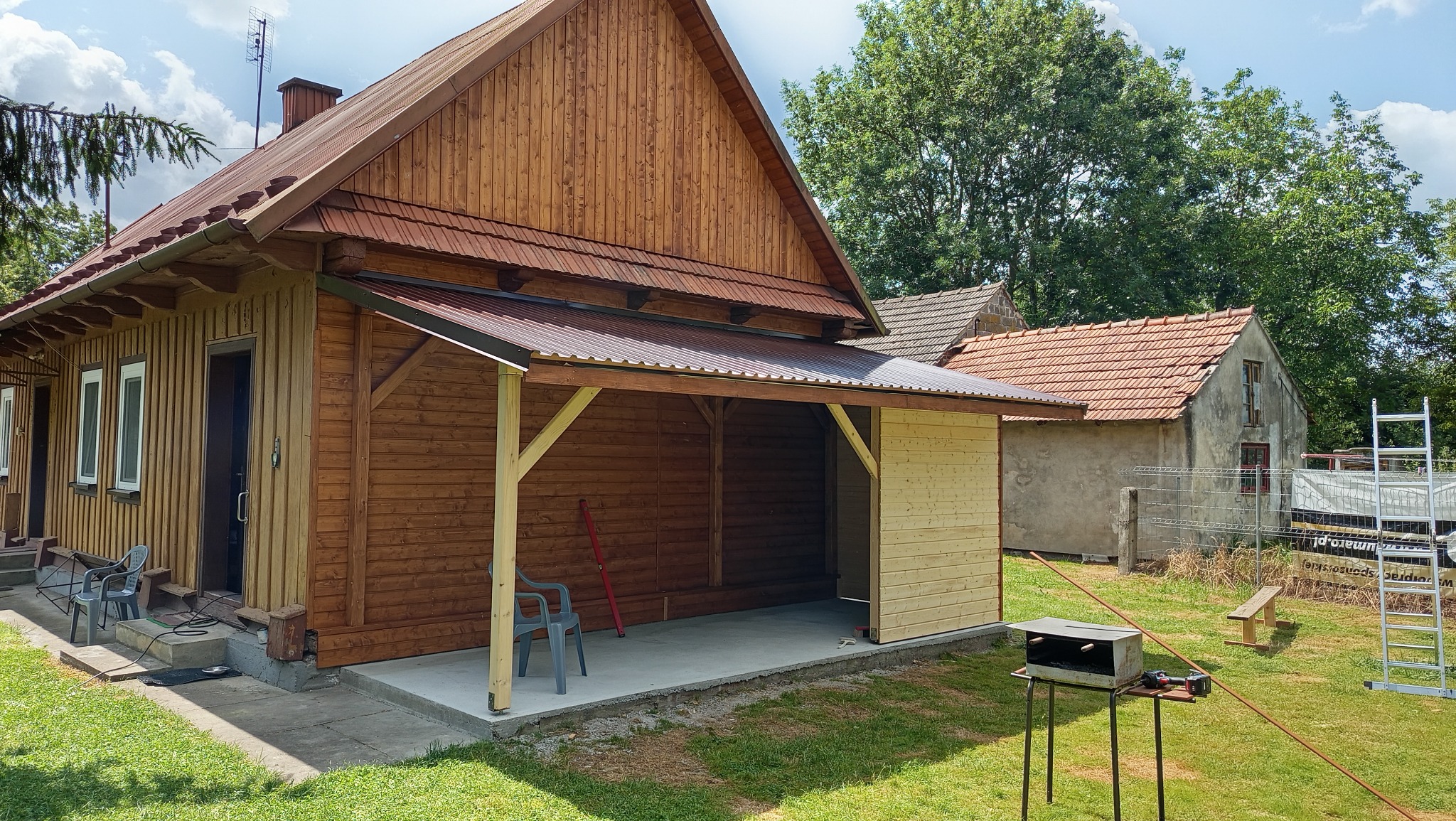 Drewniana konstrukcja z zadaszeniem przy budynku, częściowo obita deskami, z widocznym betonowym podłożem i narzędziami. W tle zieleń i budynek z czerwoną dachówką.