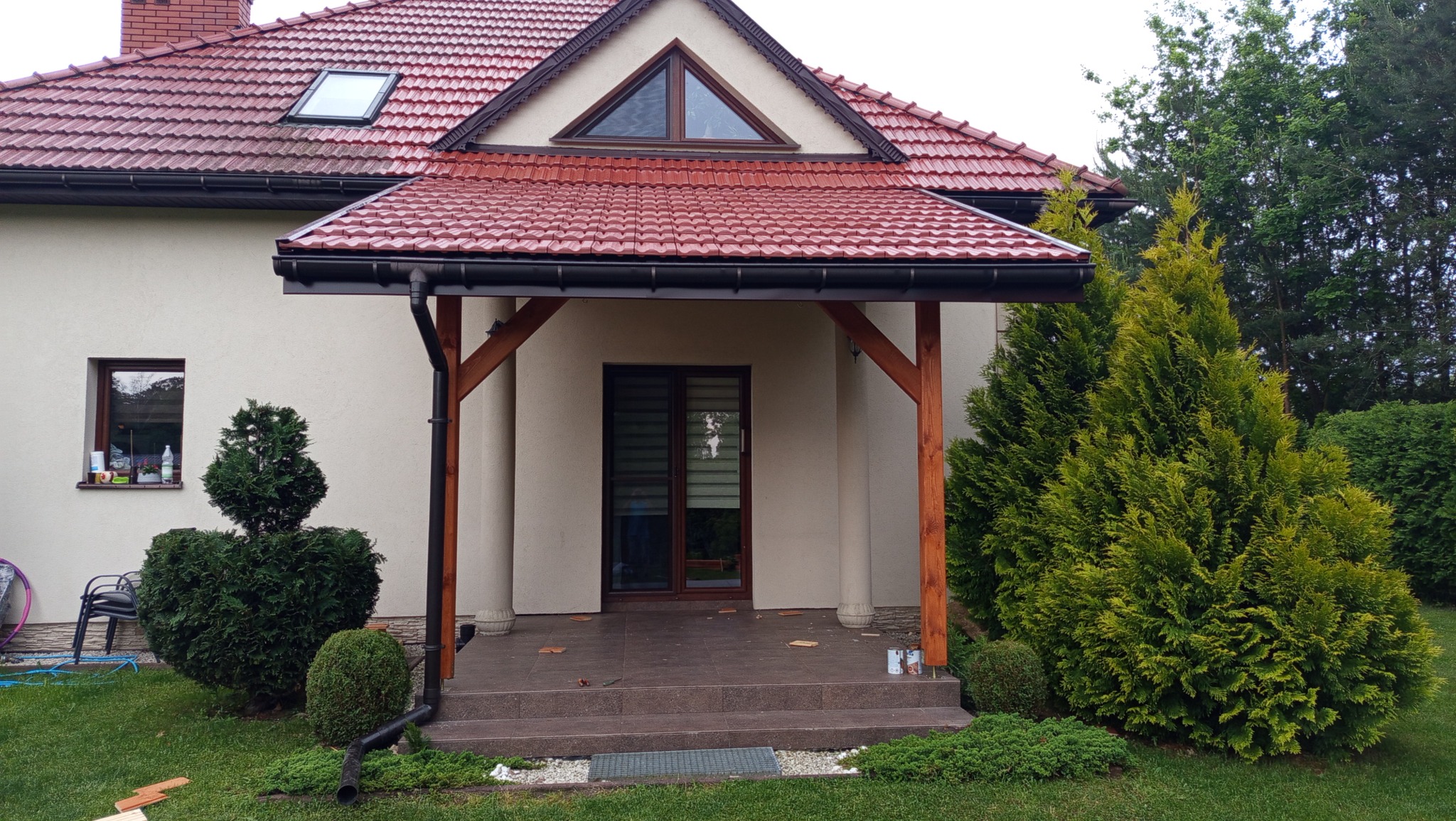 Zadaszony taras z drewnianą konstrukcją i ceramiczną dachówką, przylegający do domu z czerwonym dachem. Widoczne tuje i trawnik. Detal: ozdobne zakończenia słupów.