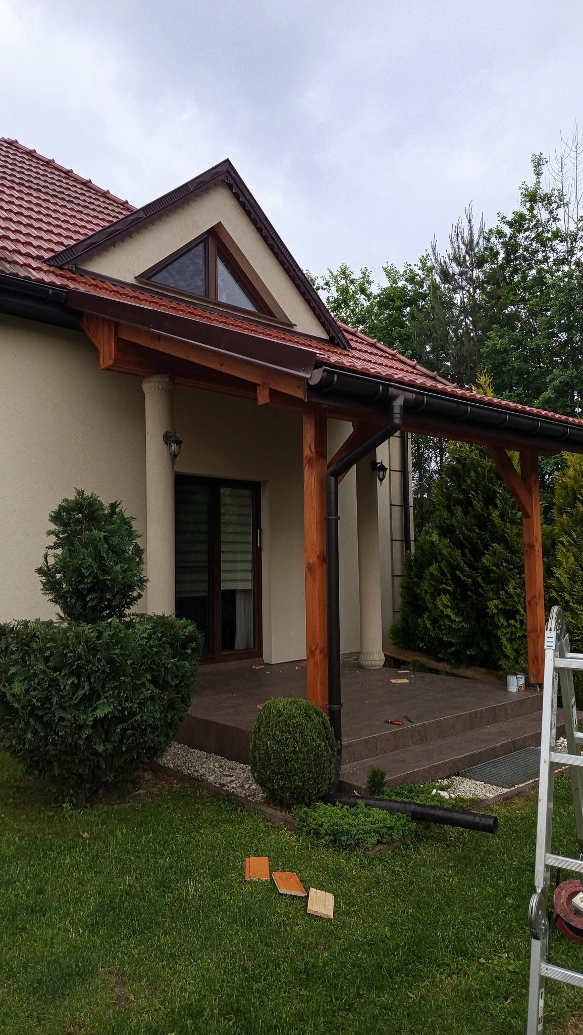 Zadaszenie tarasu z drewnianą konstrukcją i kolumnami, widok z ogrodu. Na trawniku leżą fragmenty płytek. Częściowo widoczna drabina.