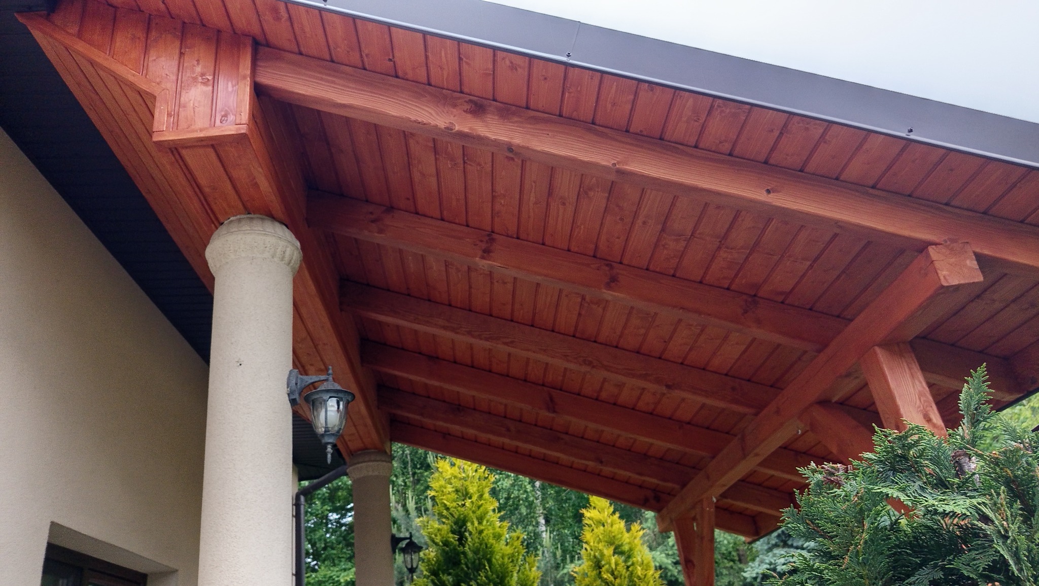 Zadaszenie tarasu z drewnianą konstrukcją i kolumną z oświetleniem, widok od dołu. Czerwono-brązowe drewno kontrastuje z zielenią drzew w tle.