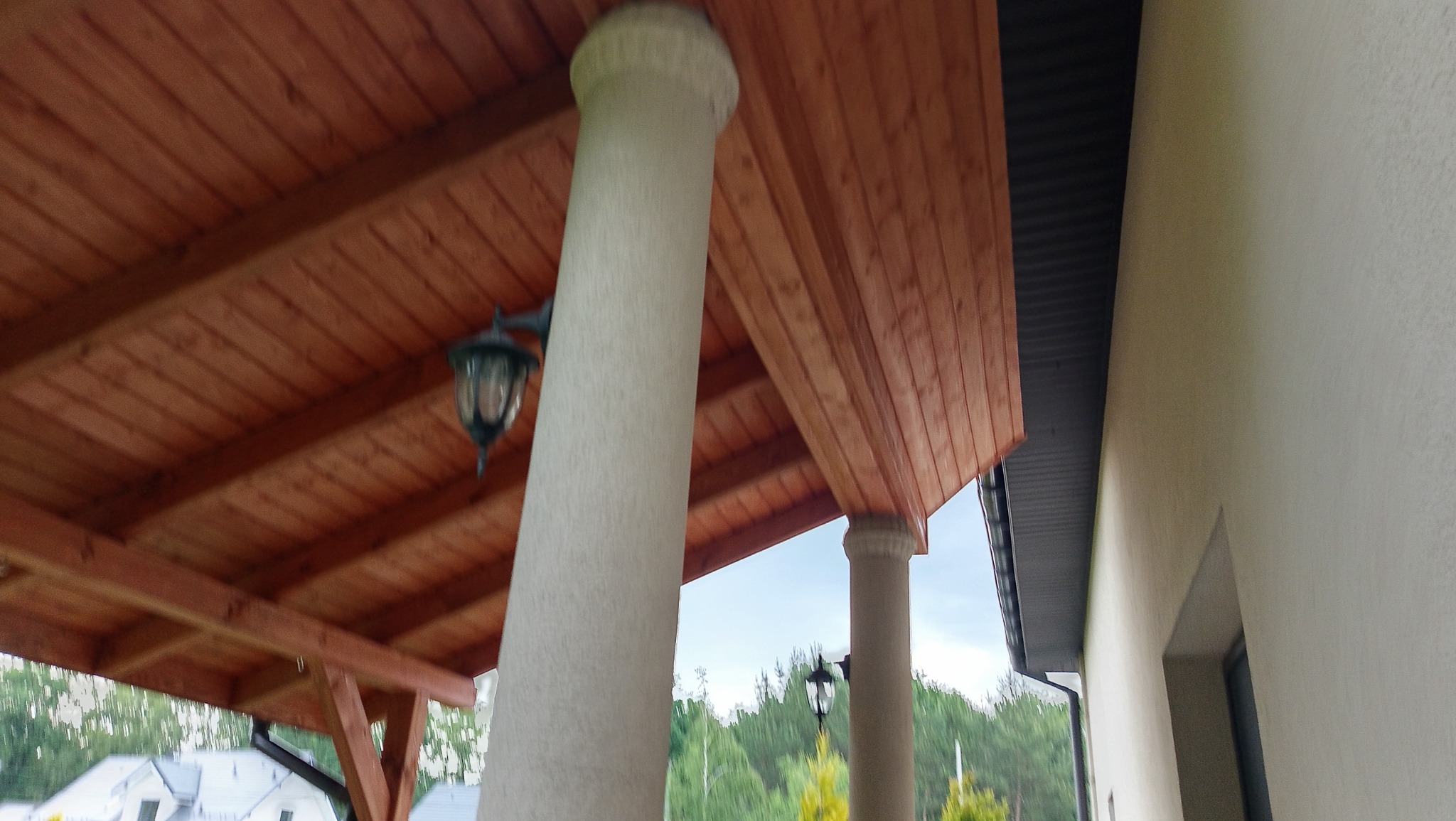 Zadaszenie tarasu z drewnianą podbitką i kolumnami oświetlonymi lampami. Widok od dołu, w tle zieleń i fragment budynku.