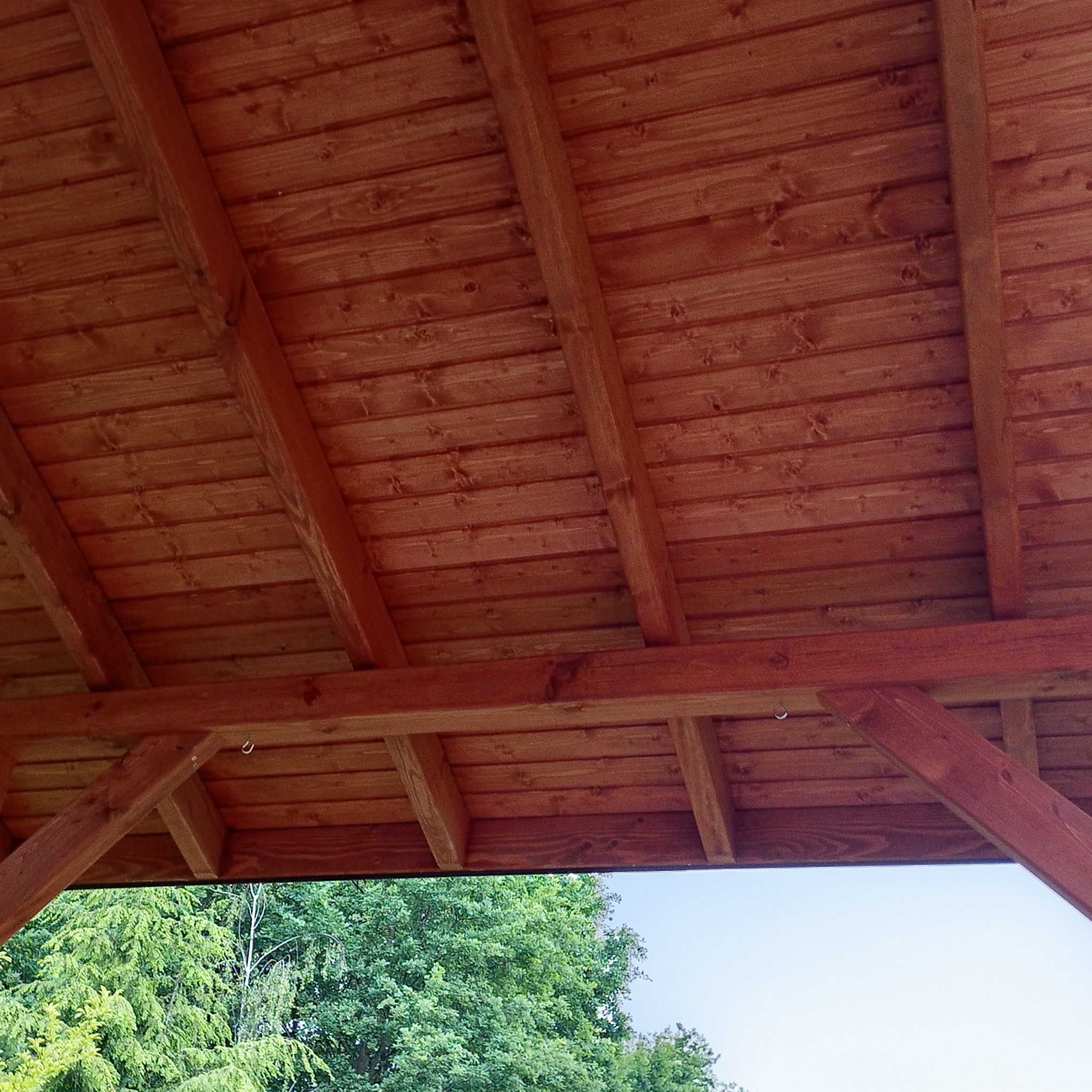 Wnętrze drewnianej konstrukcji z widocznym dachem z desek i fragmentem zieleni w tle. Widok od dołu, ukazujący detale drewnianych elementów.