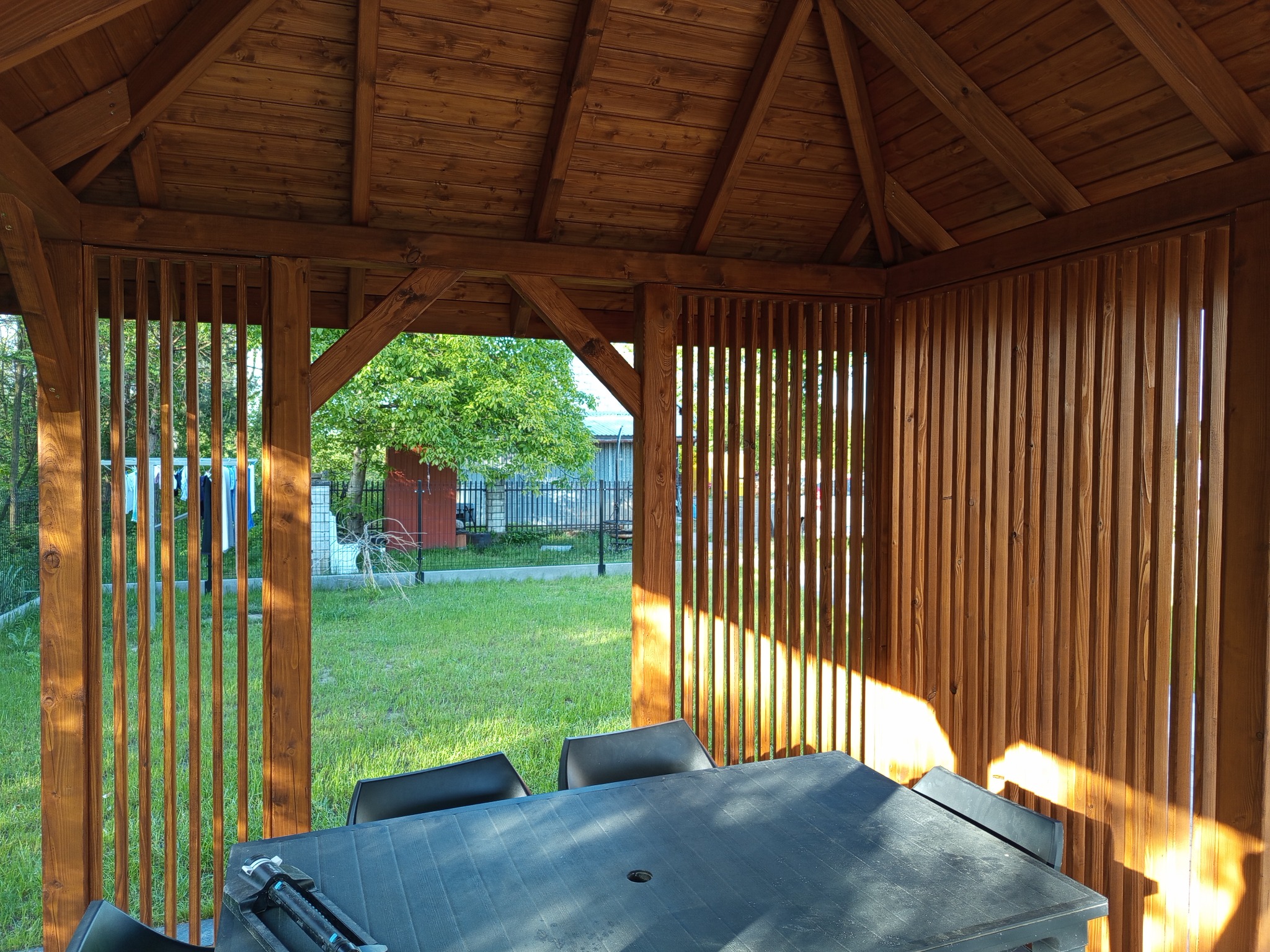 Drewniana altana z ażurowymi ścianami, wewnątrz stół i krzesła. Widok na zielony ogród przez otwarte przestrzenie. Ciepłe, naturalne oświetlenie.