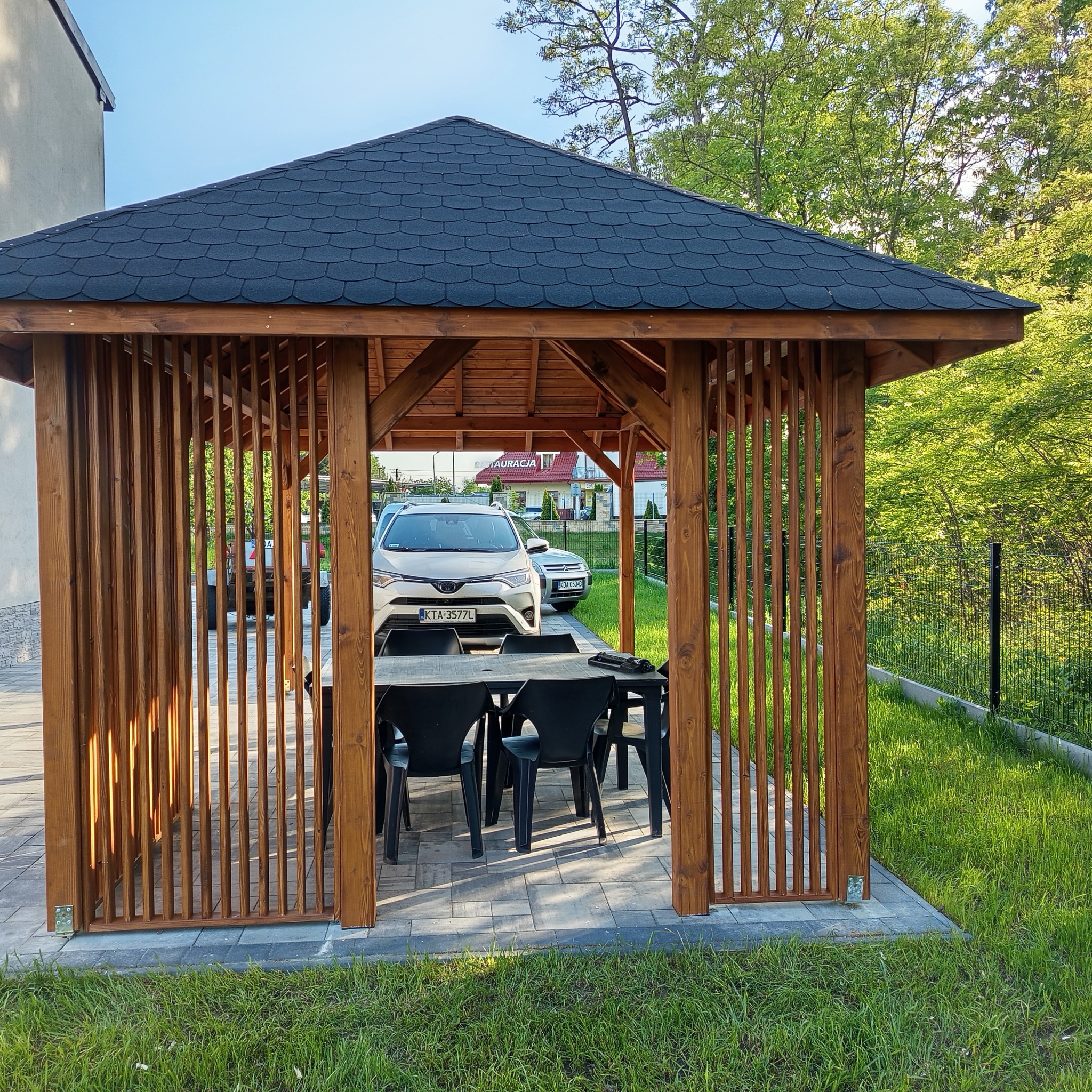 Drewniana altana z ciemnym, spadzistym dachem i ażurowymi ścianami. Wewnątrz stół z czarnymi krzesłami. W tle samochody i zieleń ogrodu. Ujęcie z poziomu gruntu.
