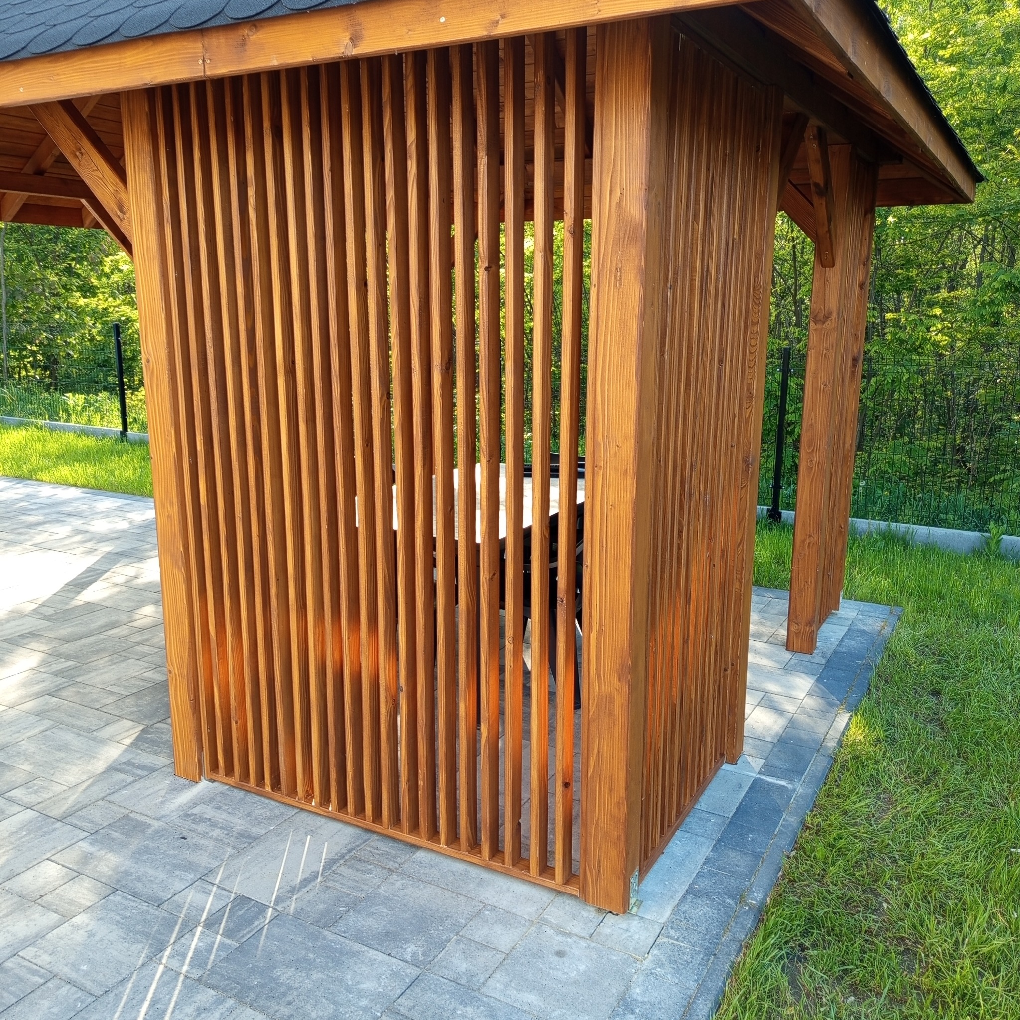 Drewniana altana z pionowymi listwami, zadaszona, na kostce brukowej. Widok na zieleń w tle. Naturalne oświetlenie, ciepłe odcienie drewna.