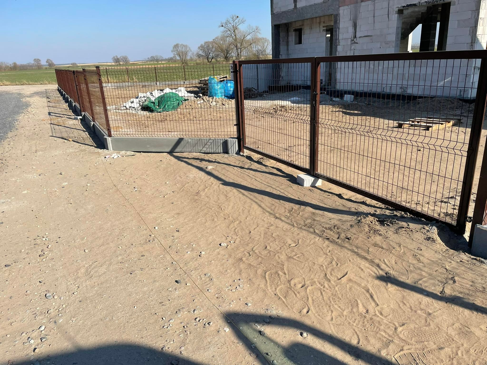 Montaż brązowej, metalowej bramy panelowej z widocznym fragmentem ogrodzenia panelowego na tle budowy domu w stanie surowym otoczonego polem.