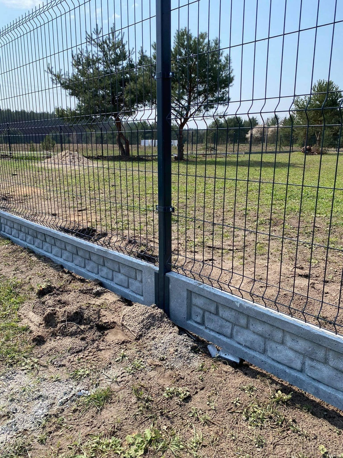 Nowo zamontowane, ciemne ogrodzenie panelowe z betonową podmurówką imitującą cegłę, widoczne z bliska na tle zielonego trawnika i drzew