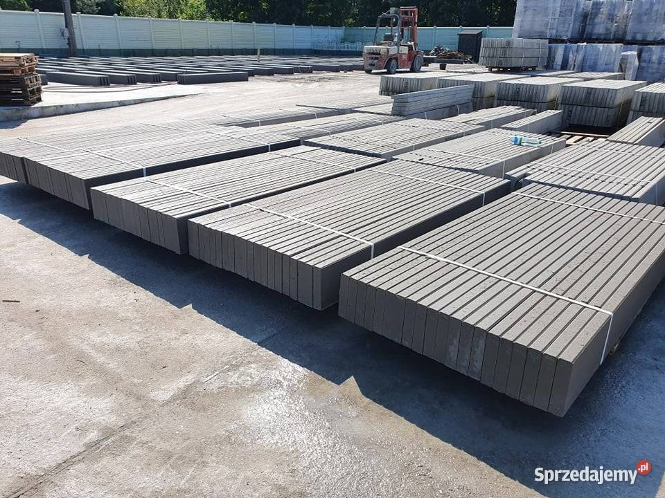 Skladowane betonowe elementy ogrodzeniowe z rowkami, spięte taśmami, na placu budowy, w tle wózek widłowy i palety z kostką brukową.