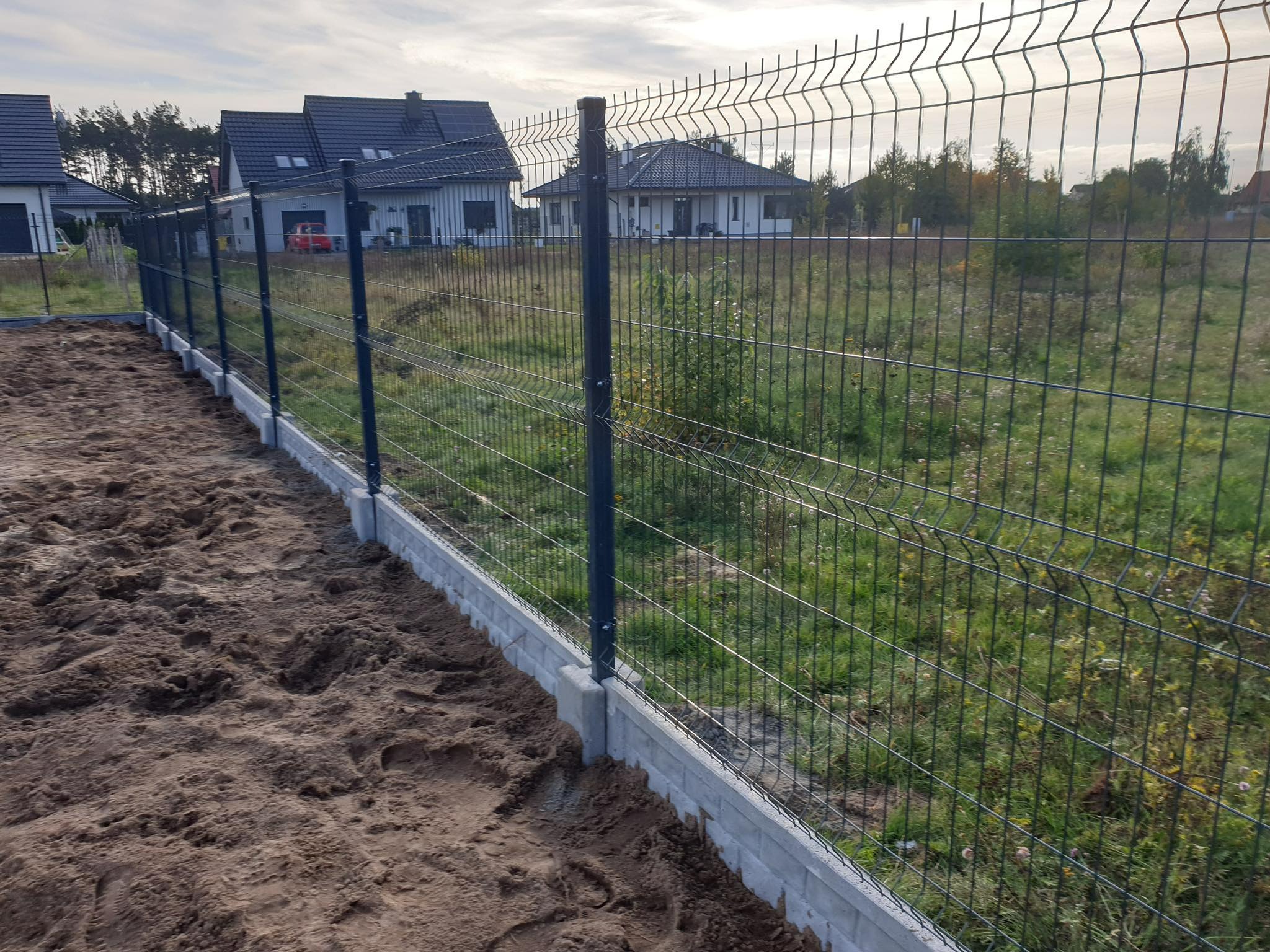 Nowo postawione, ciemnoszare ogrodzenie panelowe na betonowej podmurówce, widok na trawnik i domy w tle.