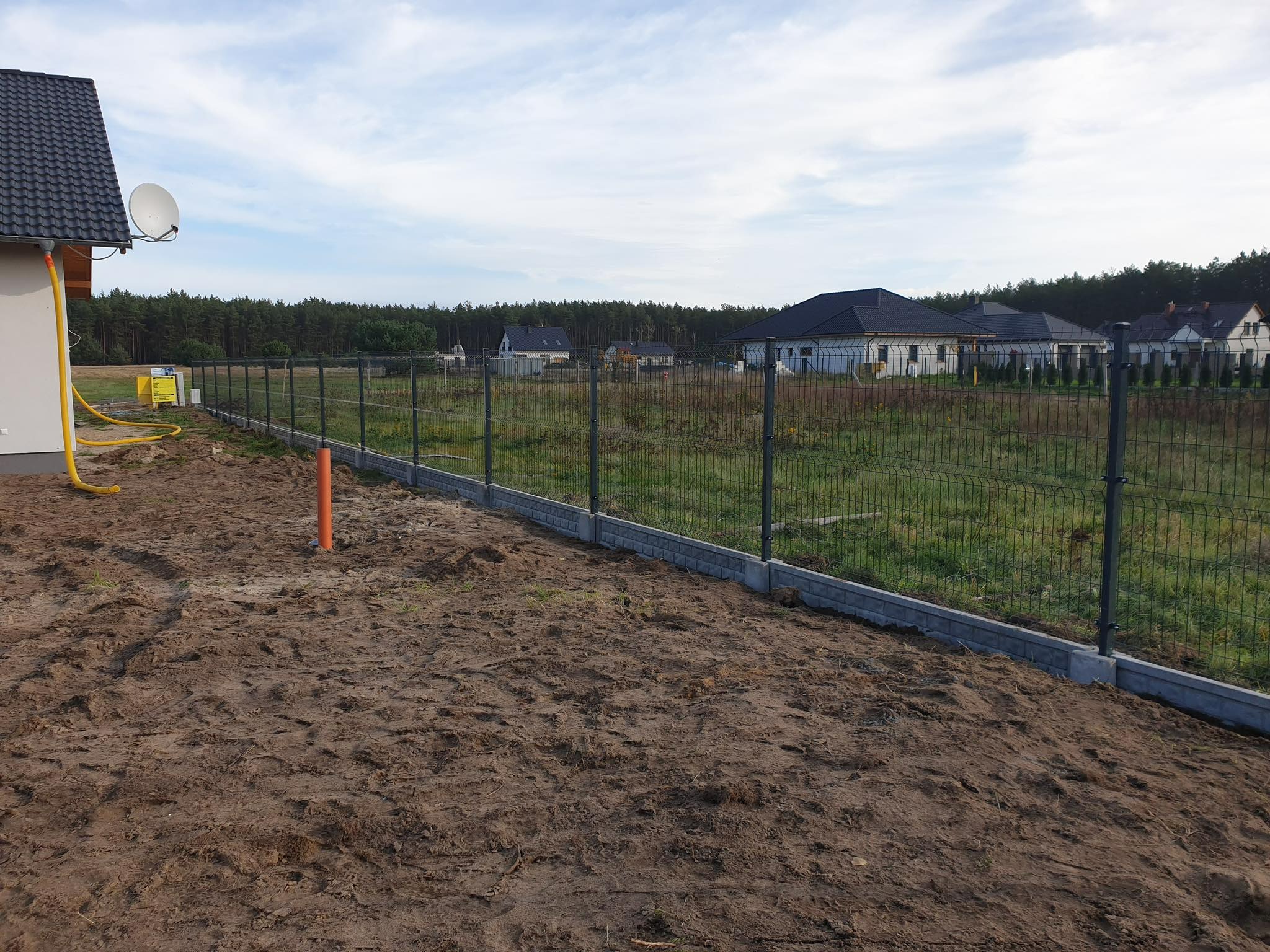 Nowo postawione, szare ogrodzenie panelowe na betonowej podmurówce, widok z bliska na brązową ziemię i fragment domu z anteną satelitarną w tle.
