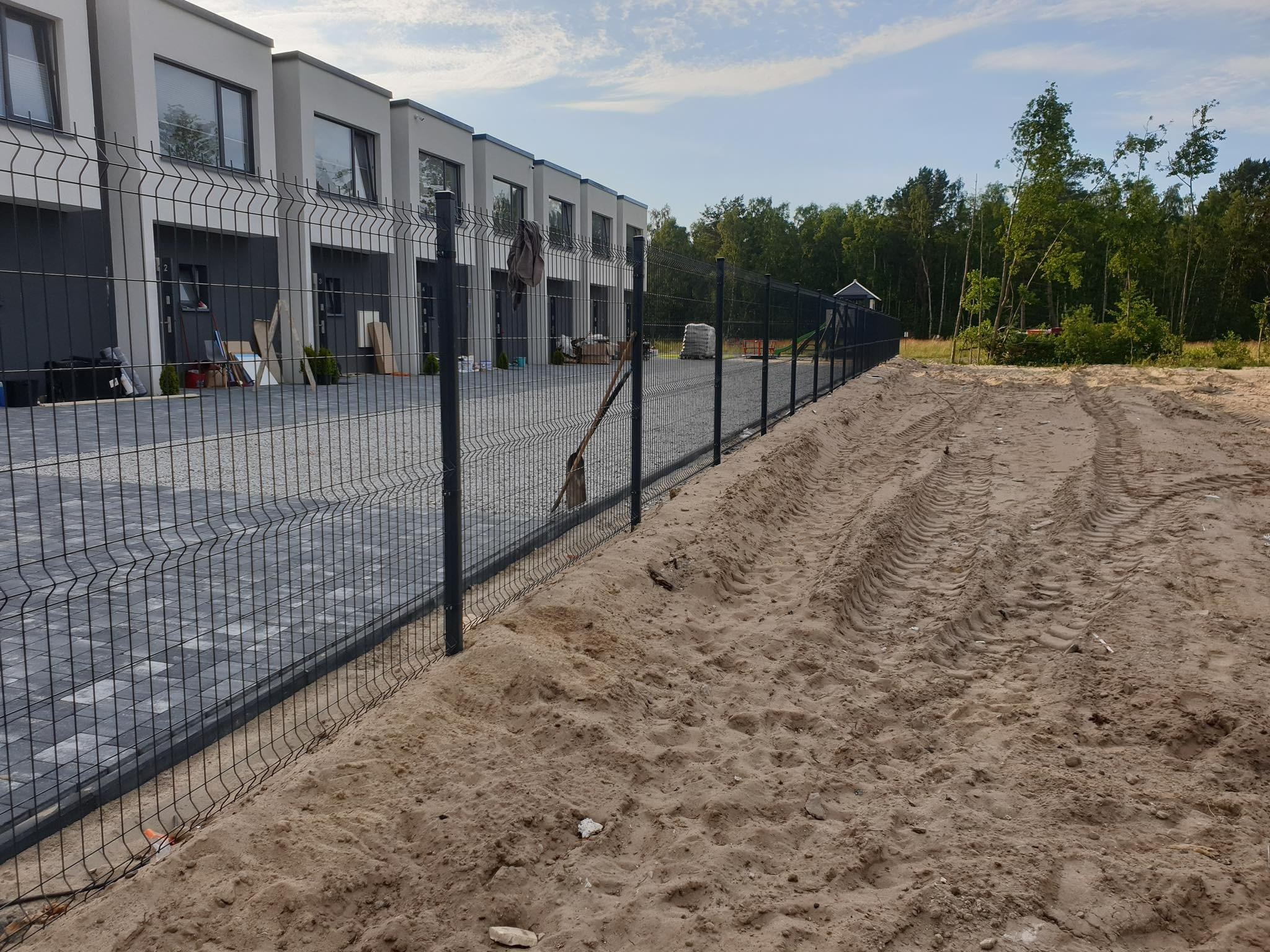 Nowoczesne, antracytowe ogrodzenie panelowe na tle szeregu domów, z piaskowym podłożem i śladami opon.