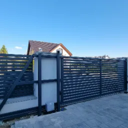 Nowoczesna, przesuwna brama wjazdowa z poziomych, szarych profili, widoczny automat do bramy, jasna kostka brukowa, domy w tle pod błękitnym niebem.