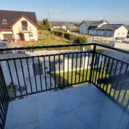 Czarna, metalowa balustrada balkonowa z widokiem na osiedle domów jednorodzinnych w słoneczny dzień.