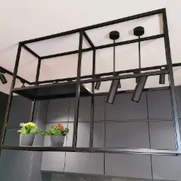 Czarna, metalowa konstrukcja sufitowa z oświetleniem punktowym i półką, na której stoją dwie doniczki z kwiatami, na tle szarych szafek kuchennych.