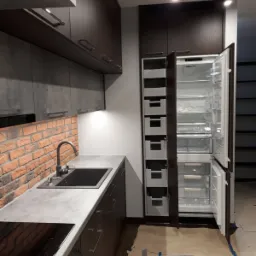 Nowoczesna kuchnia z ciemnymi, matowymi szafkami, blatem w odcieniach szarości i ceglaną ścianą, z widoczną otwartą lodówką z wewnętrznymi szufladami.