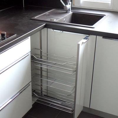 Nowoczesna kuchnia z białymi frontami, czarnym blatem i wysuwanym koszem cargo na akcesoria kuchenne.