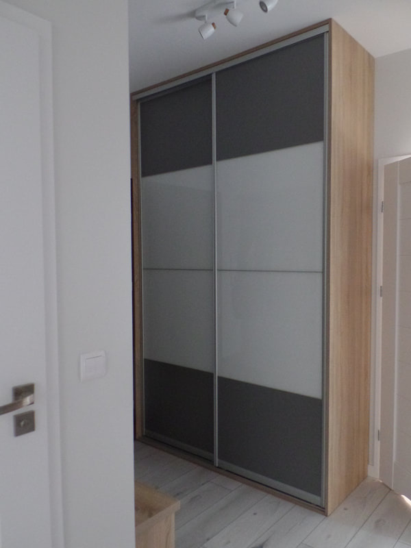 Nowoczesna szafa wnękowa z przesuwnymi drzwiami z paneli w odcieniach szarości i bieli, widoczna w jasnym wnętrzu z drewnianą podłogą.