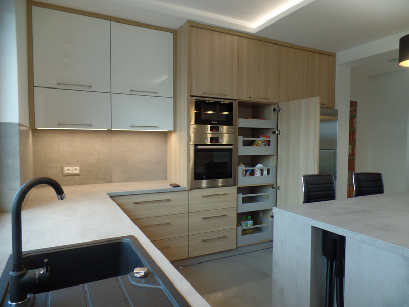 Jasna kuchnia z drewnianymi frontami i białymi szafkami wiszącymi, z otwartą szafką cargo z półkami na produkty spożywcze, widoczny czarny zlew i bateria oraz fragment blatu.