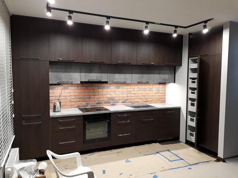 Ciemna kuchnia z drewnopodobnymi frontami, szarym blatem i ceglaną ścianą nad blatem, oświetlenie szynowe, widoczne niedokończone prace i zabezpieczenia podłogi.