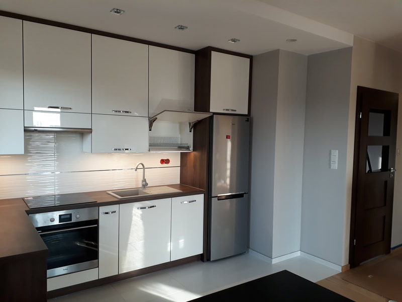 Nowoczesna kuchnia z białymi lakierowanymi frontami szafek, ciemnobrązowymi blatami i obudową, oraz srebrną lodówką w zabudowie. Widoczny reling na naczynia pod szafką wiszącą.