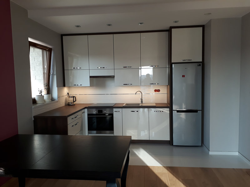 Nowoczesna kuchnia z białymi, lakierowanymi frontami szafek, ciemnobrązowym blatem i obramowaniem, oraz srebrną lodówką, widziana z perspektywy stołu jadalnego.