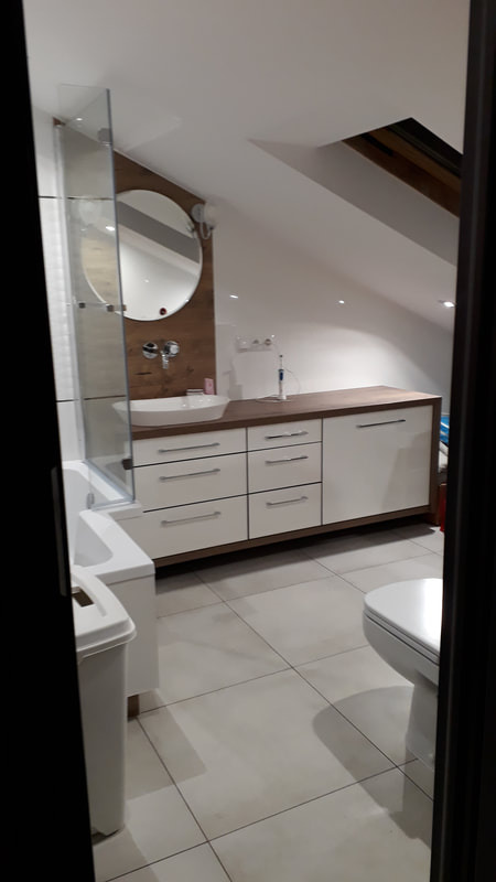 Łazienka na poddaszu ze skośnym sufitem, zabudowa meblowa z białymi frontami i drewnianym blatem, okrągłe lustro z podświetleniem, biała umywalka nablatowa.